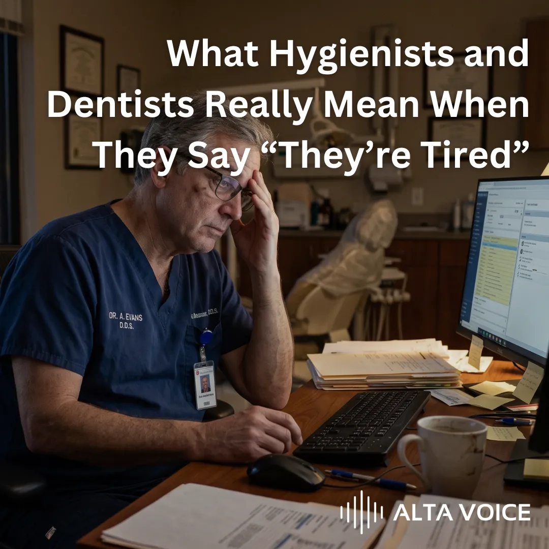 A dentist sitting at a cluttered practice computer late in the day, head in hand, with stacks of paper charts beside the keyboard.