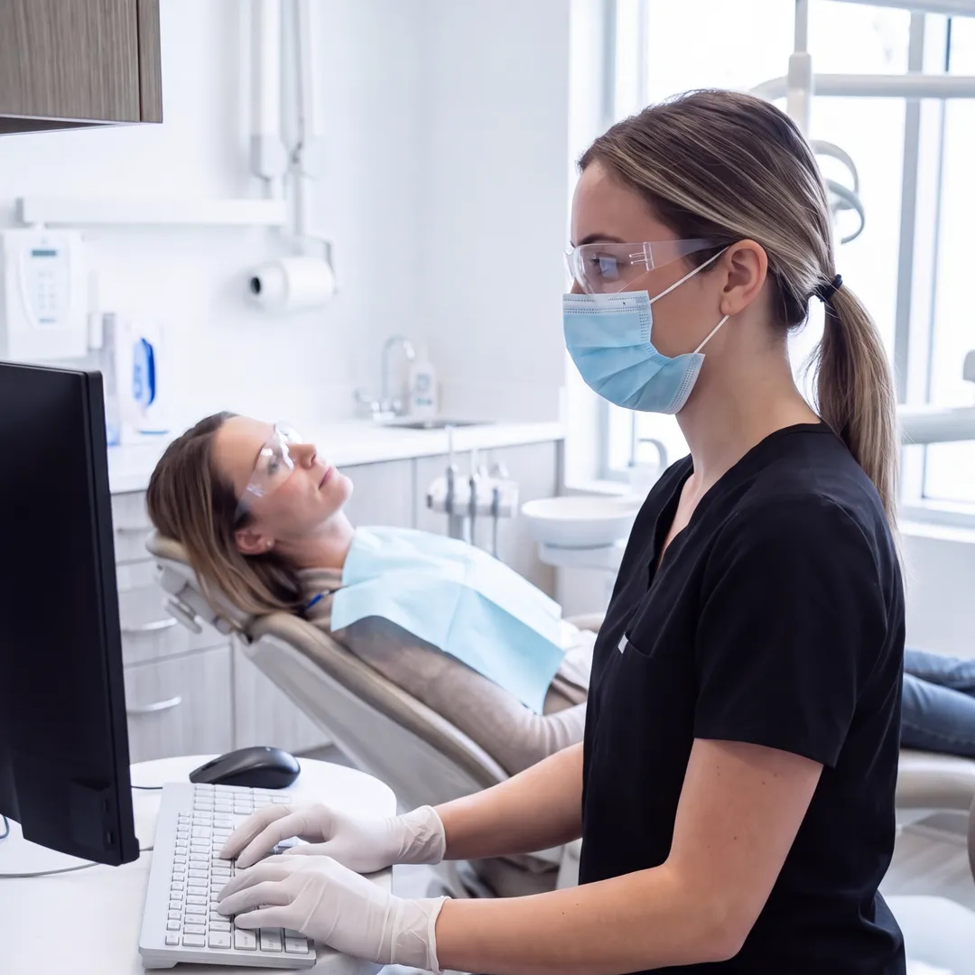 A dental hygienist in black scrubs types at the operatory computer while her patient waits in the chair behind her.